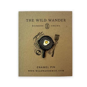 May include: Black enamel pin featuring a frying pan with a sunny-side-up egg. The pin is on a brown card with the text "THE WILD WANDER" and "RICHMOND VIRGINIA" in white. The card also says "ENAMEL PIN" and "WWW.WILDWANDERCO.COM".