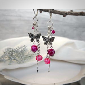 Pode incluir: Brincos pendurados com pingentes de borboleta prateados, correntes e contas rosa. Os brincos apresentam contas redondas e facetadas em tons de rosa e magenta. Os brincos são projetados com fecho de gancho.