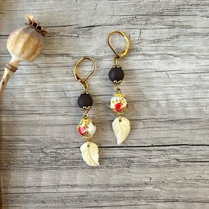 May include: Gold-coloured drop earrings with dark brown and floral beads, and ivory leaf charms. The earrings have lever-back closures. The floral beads feature red roses. The earrings are displayed on a light wood-grain surface.