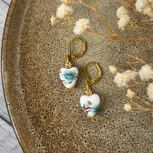 Könnte beinhalten: Ohrringe in Herzform mit floralem Design. Die Ohrringe haben eine weiße Basis mit einem blau-grünen Blumenmuster und goldfarbenen Akzenten. Sie werden auf einer braunen Oberfläche mit weißen Blumen präsentiert.