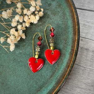 Puede incluir: Un par de pendientes en forma de corazón con un vibrante acabado de esmalte rojo. Los pendientes tienen ganchos dorados y están adornados con cuentas en tonos rojos, dorados y marrones. Los pendientes se exhiben en un plato verde azulado.