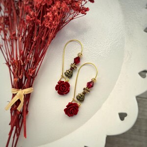 Può includere: Orecchini pendenti color oro con pendenti a forma di rosa rossa, perline rosse sfaccettate e piccoli ciondoli a forma di foglia color bronzo. Esposti su una superficie bianca con un bouquet di fiori secchi rossi.