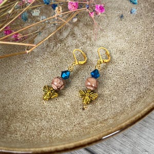 May include: A pair of gold-colored earrings with a bee charm, a pink and brown bead, and a blue crystal. The earrings are displayed on a brown plate with dried flowers in the background.