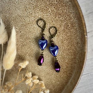 Op de afbeelding: Een paar oorhangers met hartvormige blauwe glazen kralen en druppelvormige paarse kralen. De oorbellen hebben bronskleurige oorhaken en accenten. De oorbellen worden gepresenteerd op een bruine plaat met gedroogde bloemen.