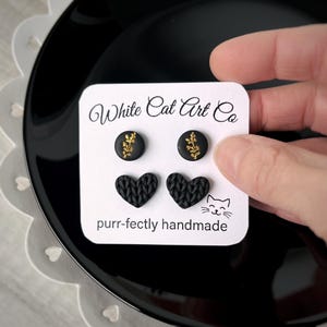 Könnte beinhalten: Schwarze Ohrstecker mit goldenen Blattdetails und schwarze herzförmige Ohrringe. Die Ohrringe werden auf einer weißen Karte mit dem Text "White Cat Art Co" und "purr-fectly handmade" präsentiert.