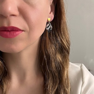 Puede incluir: Pendientes con un pendiente de botón dorado en forma de corazón y un pendiente de lágrima veteada en negro, blanco y gris. La persona lleva una blazer blanca.