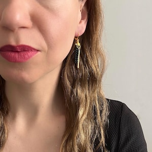May include: Gold-tone earrings with a black woven design and a vertical gold bar. The earrings are dangling from a gold hook. The earrings are a stylish accessory.