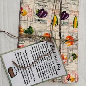 Può includere: Una borsa di stoffa beige con stampa di verdure, legata con spago, e un'etichetta che dice "Baked Potato Bag!". La borsa presenta illustrazioni di pomodori, melanzane e mais. L'etichetta fornisce istruzioni.