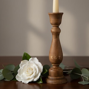 Puede incluir: Un candelabro de madera con una vela blanca encendida. El candelabro es marrón con un diseño decorativo y las iniciales "K & T" grabadas en el lateral. Una flor blanca y vegetación están en primer plano.