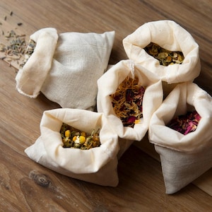 Puede incluir: Cinco pequeñas bolsas de lino abiertas llenas de hierbas y flores secas, incluyendo lavanda, manzanilla y pétalos de rosa. Las bolsas están dispuestas sobre una superficie de madera, con algunas hierbas esparcidas.