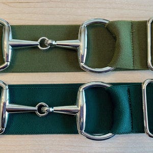 Puede incluir: Dos cinturones ecuestres verdes. El cinturón superior es verde oliva y el inferior es verde bosque oscuro. Cada cinturón tiene herrajes de bocado y hebilla plateados. Los cinturones están hechos de tela resistente.