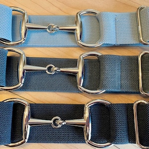 May include: Three equestrian-themed belts in shades of blue. Each belt features a silver-colored snaffle bit buckle and a rectangular metal loop. The belts are made of a woven fabric and are displayed on a wooden surface.
