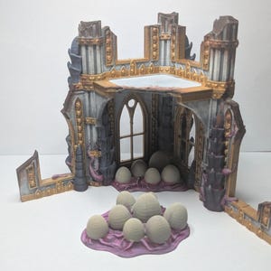 Könnte beinhalten: Ein Miniatur-Ruinenbau mit gewölbten Fenstern und aufwendigen Details, bemalt in Grau-, Gold- und Lilatönen. Eine Ansammlung hellfarbiger Eier befindet sich im und vor dem Gebäude, auf einer lila Basis.
