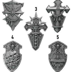 Puede incluir: Colección de cinco escudos grises detallados con varios diseños. Los escudos presentan elementos como alas, cruces, calaveras y caras. La palabra "Shields" se muestra encima de los escudos.