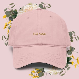 Peut inclure: Casquette de baseball rose clair avec le texte "GO MAX" brodé en or. La casquette est entourée d'un arrangement floral avec des fleurs blanches et jaunes et des feuilles vertes.