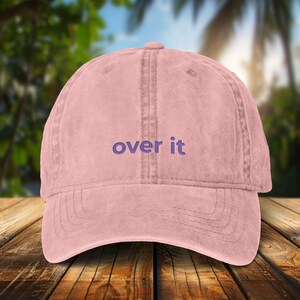 Op de afbeelding: Een roze baseballpet met de geborduurde tekst "over it" in paars. De pet heeft een gebogen klep en een gestructureerde kroon. De achtergrond is een wazige afbeelding van een tropische scène.