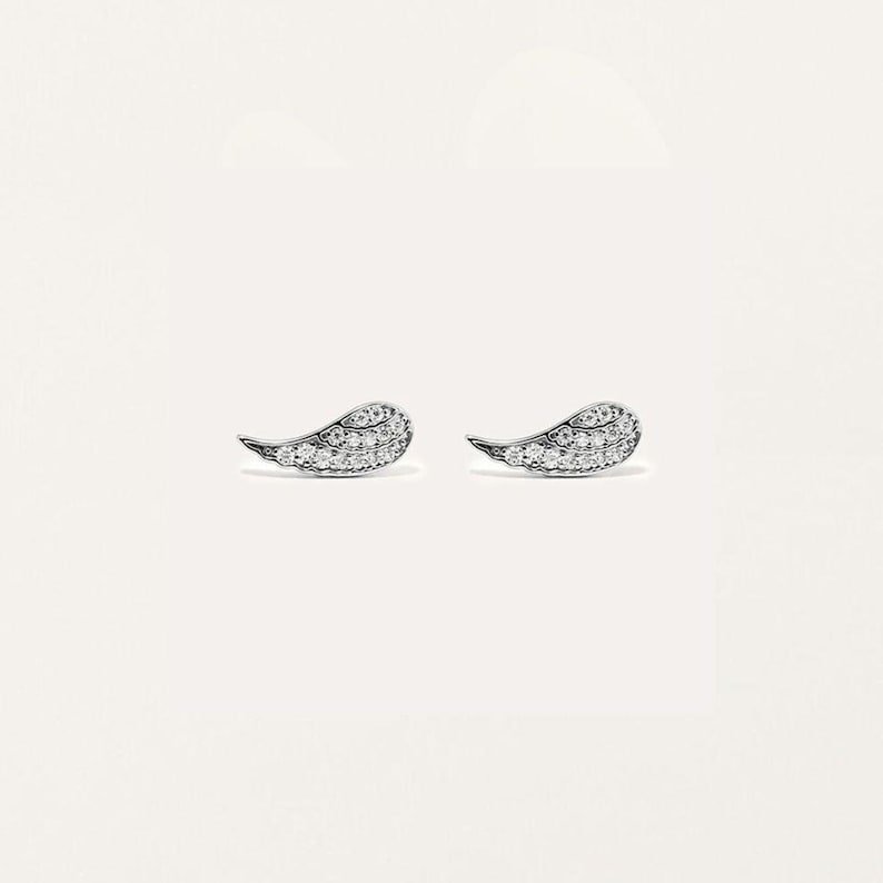 May include: A pair of silver-toned stud earrings, each shaped like a small wing. The wings are encrusted with numerous small, clear gemstones, creating a sparkling effect. The earrings are set against a plain white background.