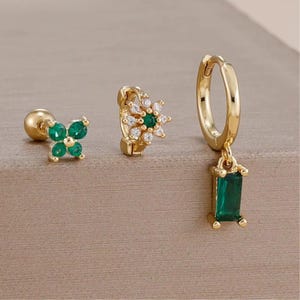 Puede incluir: Tres pendientes dorados con piedras preciosas verdes y transparentes. Uno presenta un colgante de piedra rectangular verde, otro tiene un diseño floral con piedras transparentes y el tercero un diseño de trébol con piedras verdes.