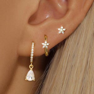 Könnte beinhalten: Ein Set aus drei goldfarbenen Ohrringen, bestehend aus einem kleinen sternförmigen Stecker mit klaren Steinen, einem kleinen Creolen-Ohrring mit Sternanhänger und klaren Steinen sowie einem Creolen-Ohrring mit einem tropfenförmigen Anhänger.