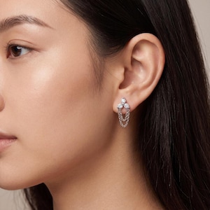 May include: A silver-toned stud earring featuring three small, clear, round-cut stones arranged in a triangular cluster at the top, with two delicate, dangling silver chains looping down from the stones to create a layered, suspended effect.