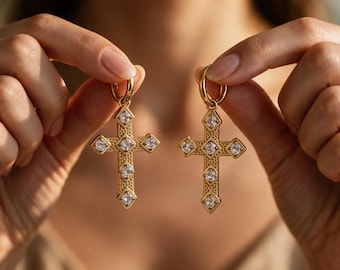 Puede incluir: Un par de colgantes de cruz en tono dorado con una superficie texturizada y granulada, adornados con cinco piedras transparentes facetadas en forma de cruz. Cada colgante está unido a una argolla dorada, mostrando un diseño de metal detallado.