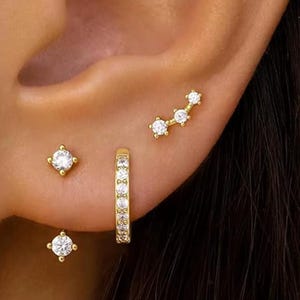 Puede incluir: Un conjunto de pendientes dorados. Hay dos pequeños pendientes de botón con piedras claras cuadradas, un pequeño pendiente de aro con múltiples piedras claras y un pendiente de barra curvada con tres piedras claras.