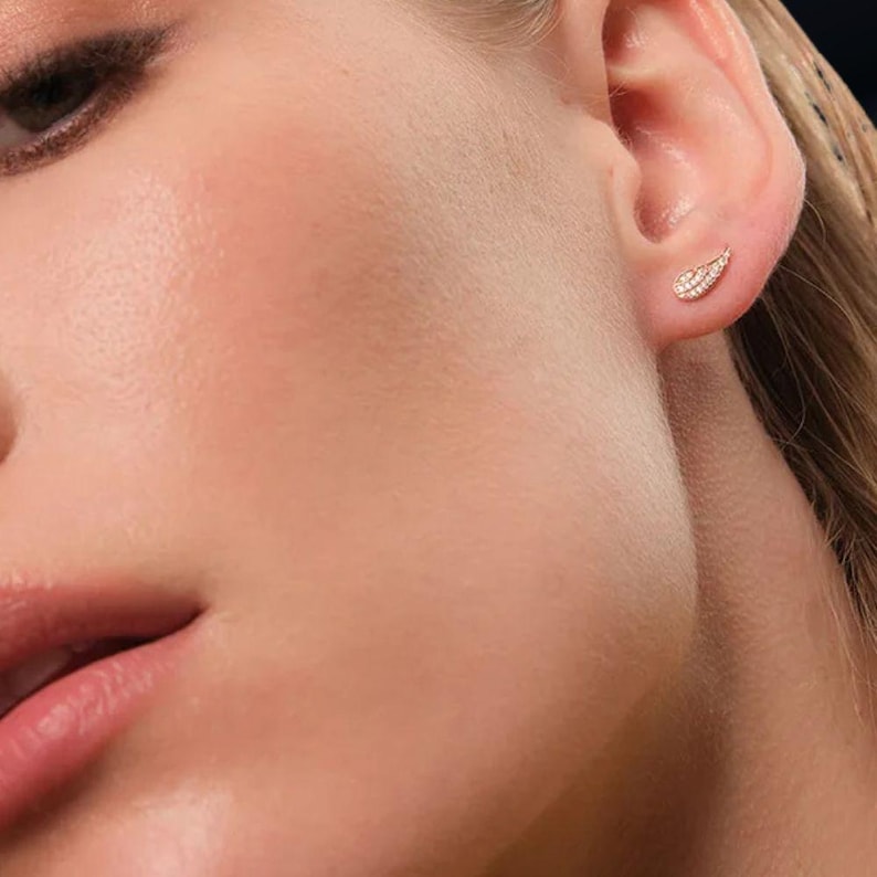 May include: Close-up of a person's ear adorned with a small, gold-toned stud earring. The earring is shaped like a leaf or feather and is encrusted with small, sparkling gemstones. The skin has a smooth, healthy appearance.