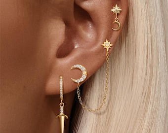 Op de afbeelding: Goudkleurige oorbellen met een hemels thema. De set bevat een dolkvormige oorbel, een halve maan oorbel, een ster oorbel en een ketting die een ster met een maan verbindt. De oorbellen zijn versierd met kleine, sprankelende steentjes.