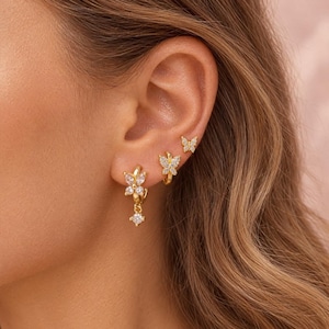 Könnte beinhalten: Drei goldfarbene Ohrringe an einem Ohr. Der obere Ohrring ist ein kleiner Schmetterlingsstecker mit klaren Steinen. Der mittlere ist ein kleiner Creolen-Ohrring mit Schmetterlingsanhänger. Der untere ist ein Creolen-Ohrring mit Schmetterling und einem hängenden klaren Stein.