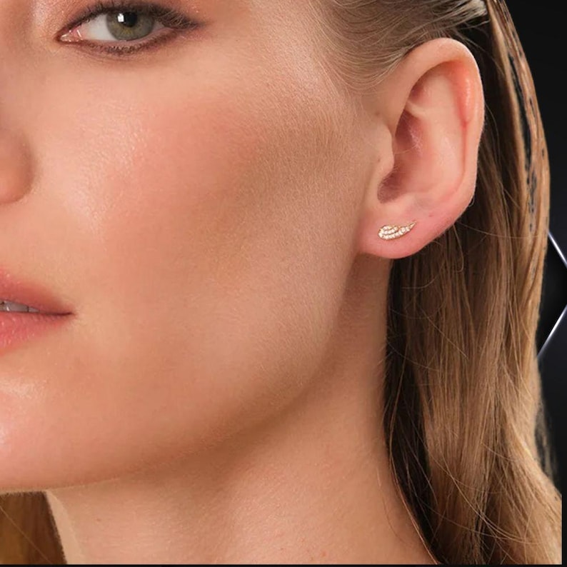 May include: Close-up of a person's ear with a small, gold-colored stud earring. The earring is shaped like a small wing and is encrusted with tiny, sparkling stones. The skin is fair with a soft, natural glow.