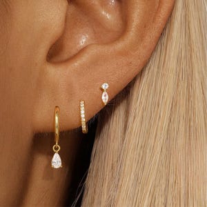 May include: A collection of gold earrings, including a small hoop with a teardrop charm, a small hoop with embedded crystals, and a stud earring with a crystal. The earrings are displayed on a person's ear.