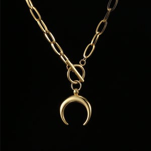 Puede incluir: Collar dorado con un colgante de media luna. El collar presenta un cierre de palanca y una cadena con eslabones ovalados. El colgante tiene forma de media luna pulida y curvada. El fondo es negro.