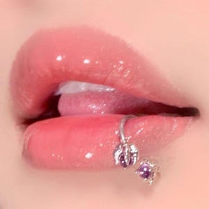 May include: Close-up of glossy, pink lips with a silver lip ring and a matching stud earring. The lip ring features a decorative design with a purple gemstone. The skin around the lips is smooth and lightly shaded.