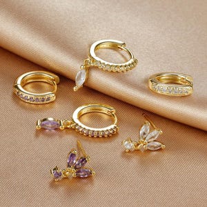 Può includere: Una collezione di orecchini dorati, tra cui modelli a cerchio e a bottone, adornati con pietre preziose chiare e viola. Gli orecchini sono esposti su una superficie di raso dorato, mettendo in risalto i loro dettagli intricati e scintillanti.