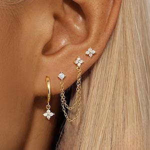 Op de afbeelding: Een verzameling goudkleurige oorbellen met heldere, sprankelende accenten. De set bevat een kleine oorring met een bungelende bloemenbedel, een oorsteker en een dubbele kettingoorbel met een bloemenbedel. De oorbellen worden op een oor getoond.