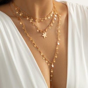 Puede incluir: Collar dorado de múltiples capas con colgantes en forma de estrella. El collar tiene tres hilos de diferentes longitudes, cada uno decorado con pequeños abalorios de estrella. El diseño es delicado y se muestra sobre un fondo blanco.