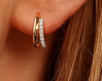 May include: Gold-tone hoop earrings with a double-band design. One earring features a curved row of small, clear gemstones. The earrings are shown on an ear, with the focus on the jewelry's details and shine.