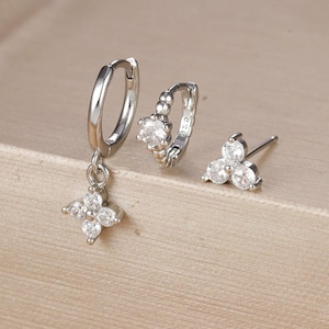 May include: A set of three silver-toned earrings. One earring is a hoop with a single clear stone, another is a stud with three clear stones, and the third is a hoop with a dangling four-leaf clover design with clear stones.
