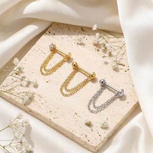 May include: Three barbell earrings featuring double dangling chains and small clear stones at each end. Two earrings are gold-toned, and one is silver-toned. Each piece consists of a straight post with a ball backing and a decorative chain link.