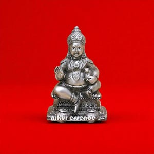 Może przedstawiać: Srebrna statua siedzącego bóstwa z bogatą biżuterią i koroną. Postać trzyma mały przedmiot i siedzi na ozdobnej podstawie. Tło jest w jednolitym czerwonym kolorze. Na podstawie widnieje napis "aikur essence".