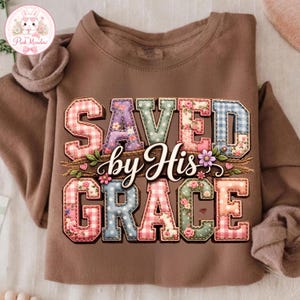 May include: A brown sweatshirt featuring the phrase "SAVED by His GRACE". The text is in a patchwork style with floral and gingham patterns. The design incorporates floral accents and wheat stalks, creating a decorative look.