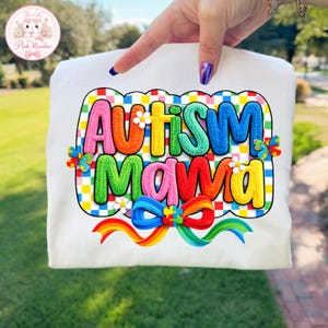 Puede incluir: Camiseta blanca con las palabras "Autism Mama" en letras coloridas y texturizadas. El diseño incluye una cinta de arcoíris, detalles de piezas de rompecabezas y un fondo a cuadros. La camiseta es sostenida por una mano.