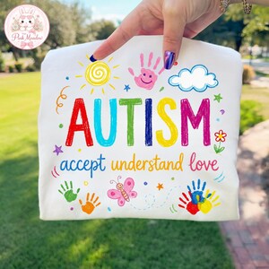 Puede incluir: Camiseta blanca con la palabra "AUTISMO" en colores arcoíris, junto con las frases "aceptar comprender amar". La camiseta presenta huellas de manos coloridas, un sol, una nube, una mariposa y estrellas. El logo de Pink Meadow está en la esquina superior izquierda.