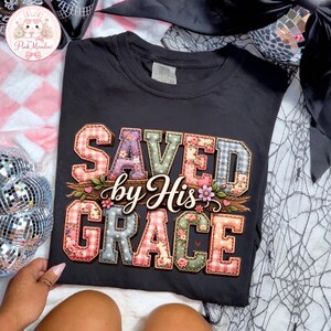 May include: Black t-shirt featuring the phrase "SAVED by His GRACE" in a patchwork design. The letters are in various colours and patterns, including floral and plaid. The shirt has a crew neck and short sleeves.