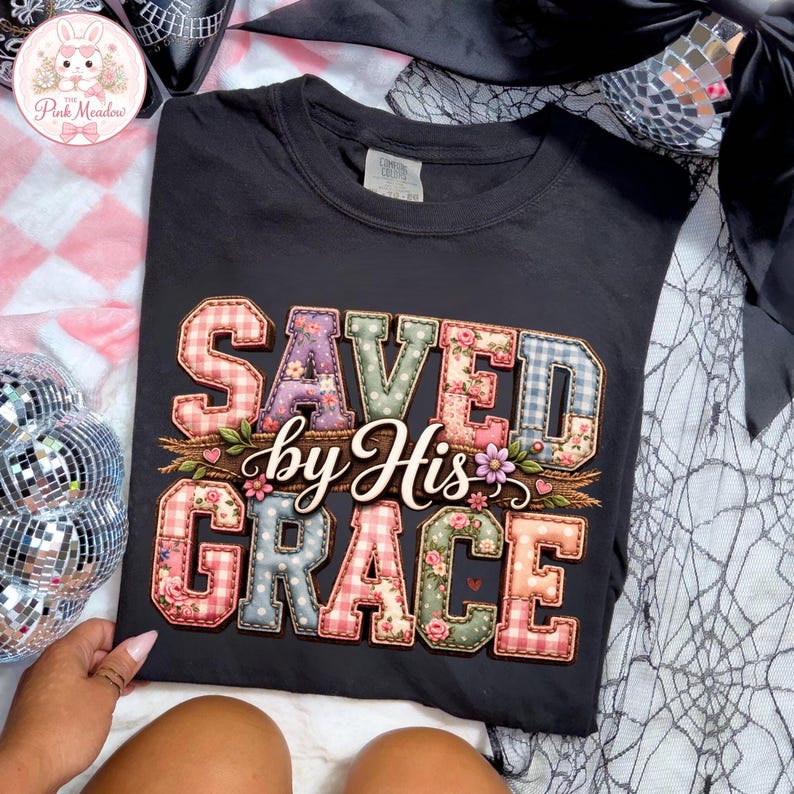 May include: Black t-shirt featuring the phrase "SAVED by His GRACE" in a patchwork design. The letters are in various colours and patterns, including floral and plaid. The shirt has a crew neck and short sleeves.