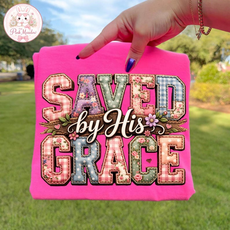 May include: A vibrant pink t-shirt with the text "SAVED by His GRACE". The letters are designed with floral and gingham patterns. The Pink Meadow logo is in the upper left corner. The shirt is a bright, eye-catching piece.