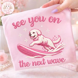 May include: Pink embroidered fabric with a surfing dog design. The text "see you on the next wave" is embroidered in a matching pink thread. The dog is white with pink accents, riding a pink surfboard.