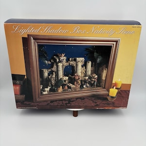 Könnte beinhalten: Eine beleuchtete Schattenbox-Weihnachtskrippe. Die Szene ist in einem dunklen Holzton gerahmt und zeigt eine detaillierte Darstellung der Geburt Christi mit Figuren, Säulen und Palmen. Die Box hat einen dunkelblauen Hintergrund mit Sternen und zwei gelben Kerzen.