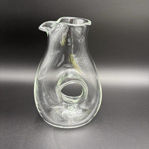 Puede incluir: Una jarra de vidrio transparente con un diseño único. Tiene un pico para verter y una gran abertura circular en el centro. El vidrio es liso y transparente. La jarra está sobre una superficie oscura.