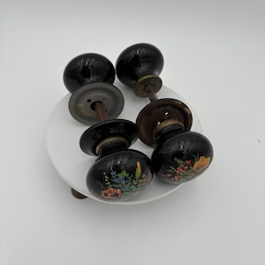 Puede incluir: Juego de seis pomos de puerta vintage de cerámica negra. Dos pomos son lisos, mientras que los otros presentan diseños florales pintados a mano en tonos naranja, azul y amarillo. Los pomos están montados sobre herrajes metálicos envejecidos.